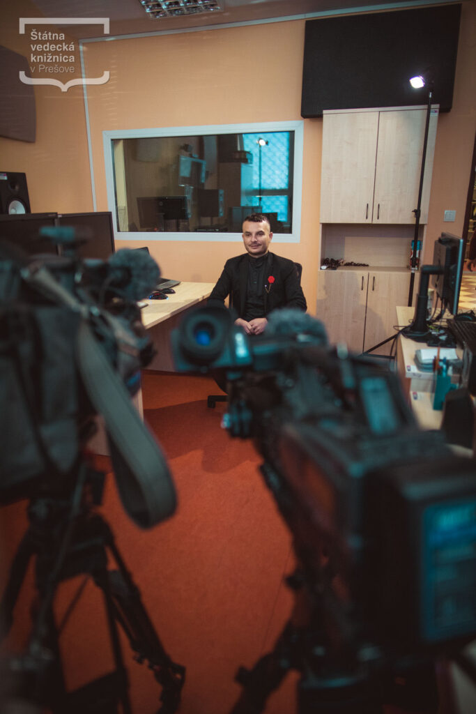 Osoba sedí v nahrávacom štúdiu počas natáčania rozhovoru, v popredí sú profesionálne videokamery a štúdiové osvetlenie.