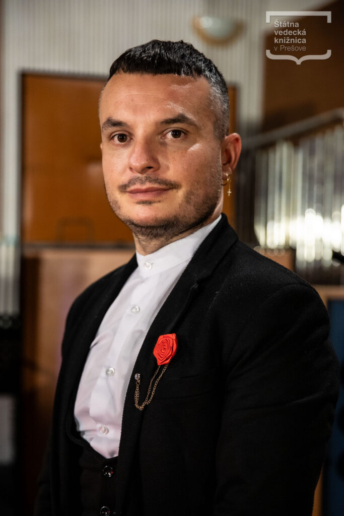 Portrét osoby v tmavom saku a bielej košeli stojacej v interiéri.
