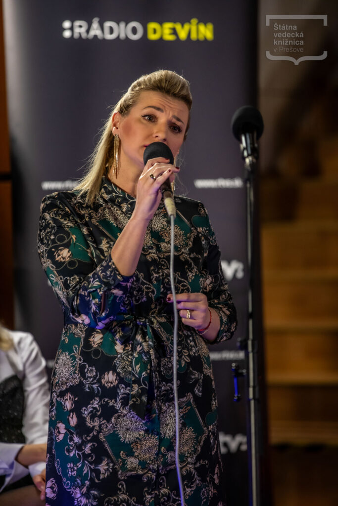 Osoba spieva do mikrofónu počas hudobného vystúpenia, v pozadí je viditeľný banner Rádia Devín.