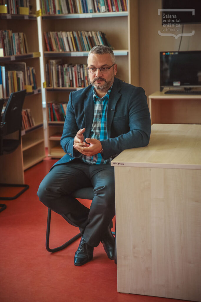 Osoba sedí v knižnici počas rozhovoru a gestikuluje rukami, v pozadí sú knižné regály.