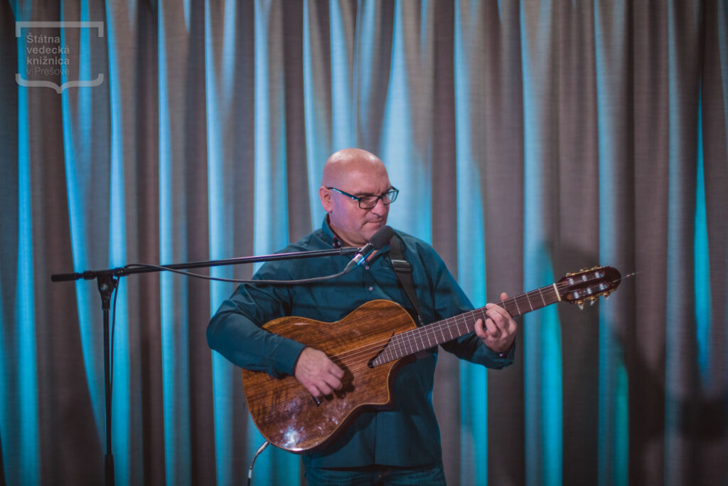 Osoba hrá na akustickej gitare počas hudobného vystúpenia na pódiu pred závesom a mikrofónovým stojanom.