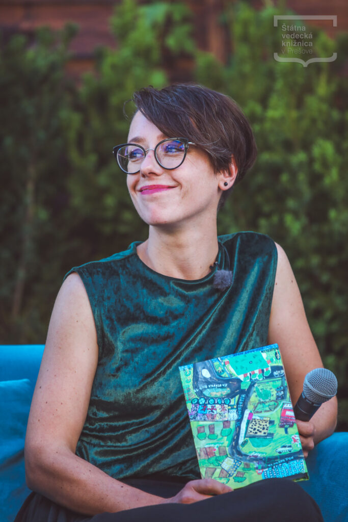 Osoba s krátkymi vlasmi a okuliarmi sedí v exteriéri, drží mikrofón a publikáciu s farebnou ilustráciou na obálke.