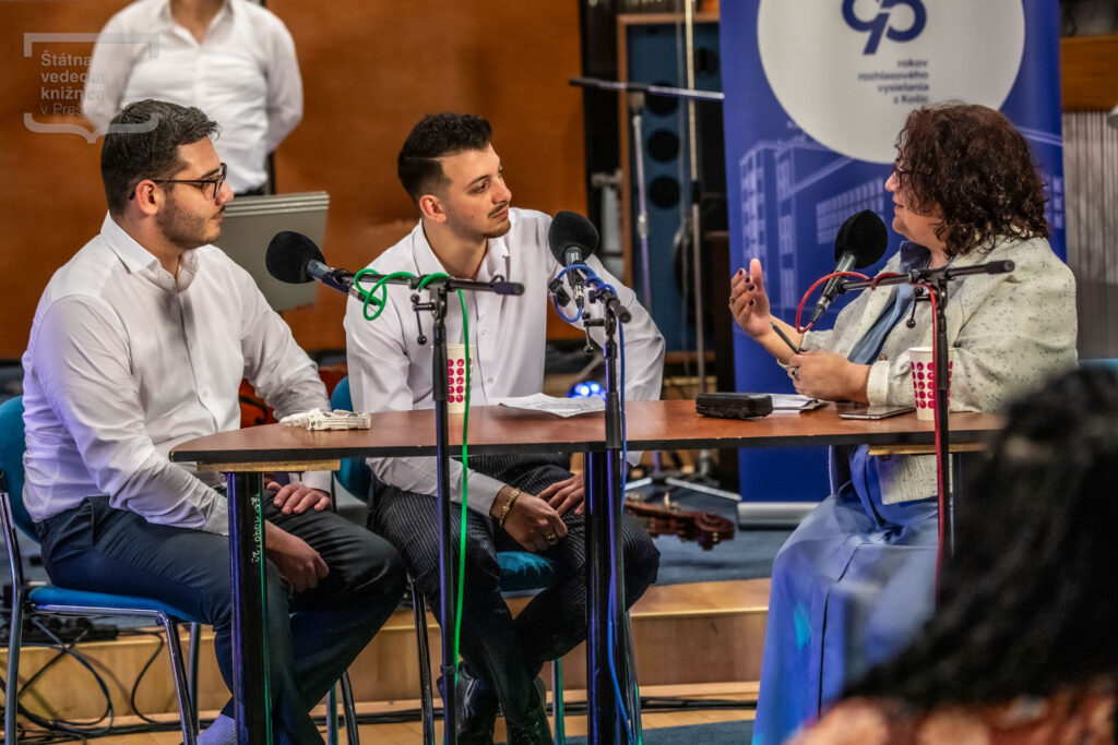 Tri osoby sedia pri stole s mikrofónmi počas diskusie na pódiu v koncertnej sále.