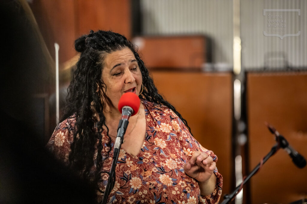 Osoba spieva do mikrofónu počas hudobného vystúpenia v koncertnom priestore.