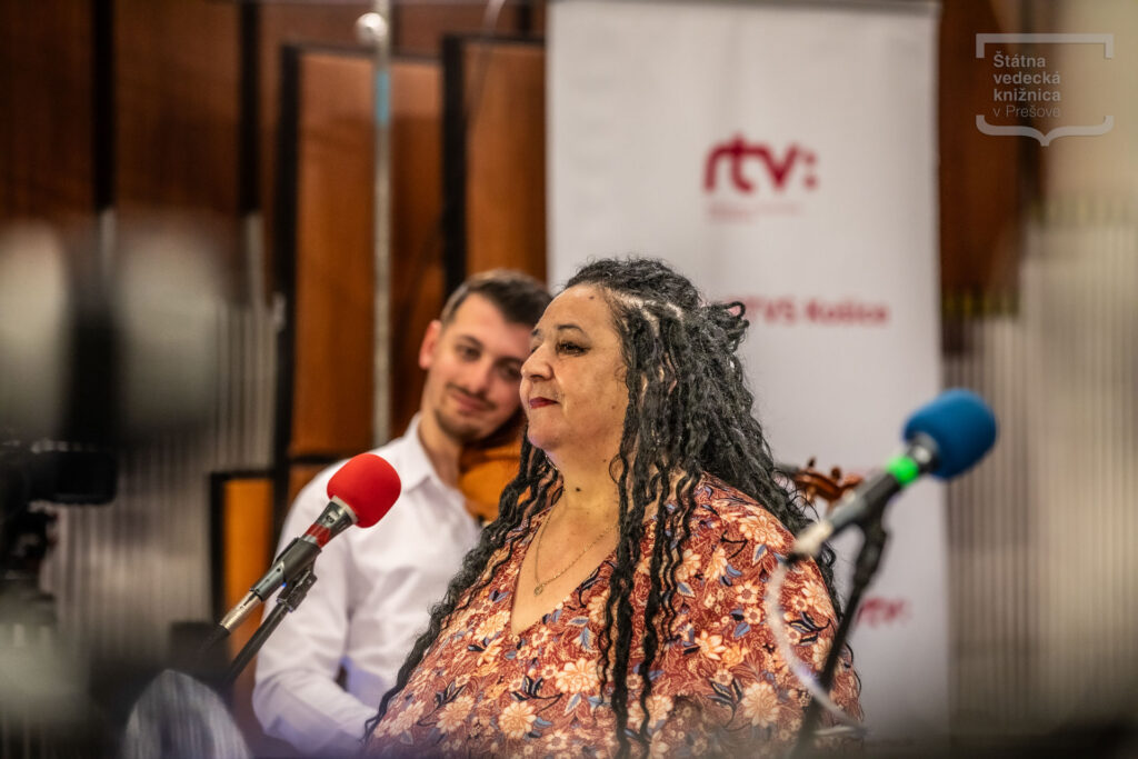 Osoba hovorí do mikrofónu počas verejnej diskusie alebo hudobného vystúpenia, v pozadí sú ďalší účastníci podujatia.