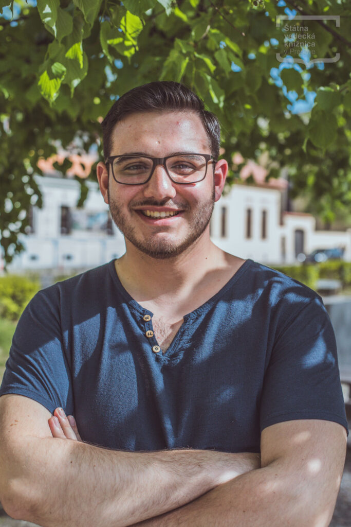 Osoba v tmavomodrom tričku a okuliaroch stojí pod stromom s prekríženými rukami.
