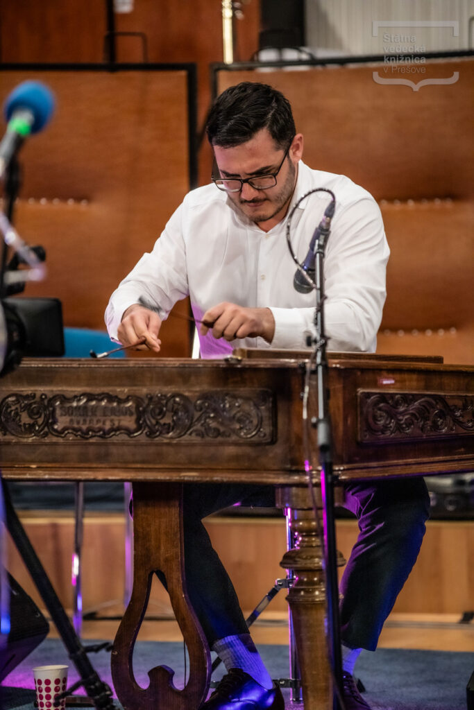 Osoba sedí za cimbalom a hrá počas koncertného vystúpenia v interiéri.