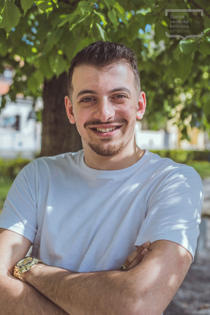 Osoba v bielom tričku stojí pod stromom v parku s prekríženými rukami.